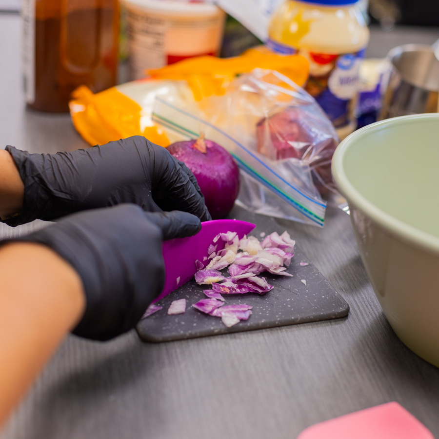 Close-up of a student in black gloves finely chopping a red onion with a purple knife on a cutting board