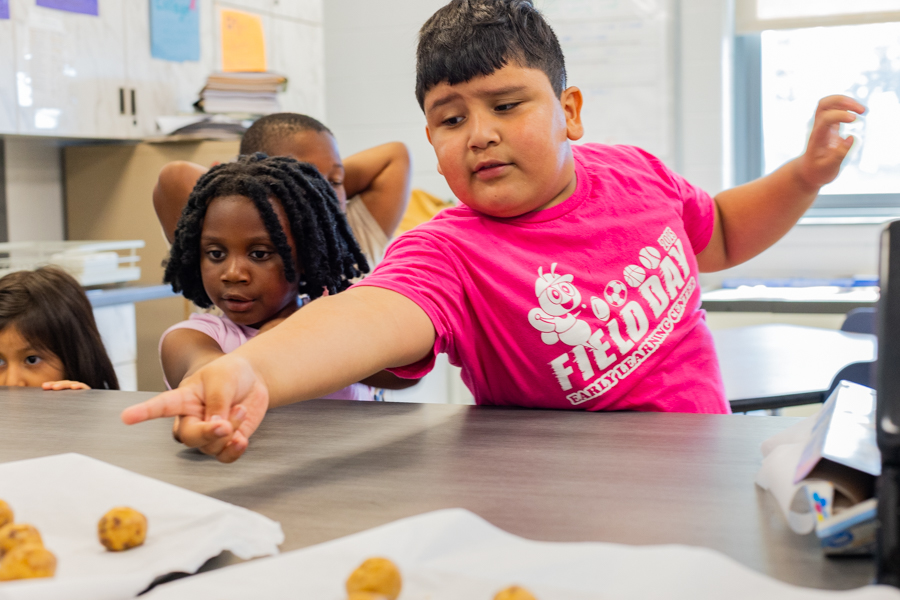 A young boy in a pink Field Day t-shirt pointing eagerly at cookie dough balls on a tray, two younger children beside him