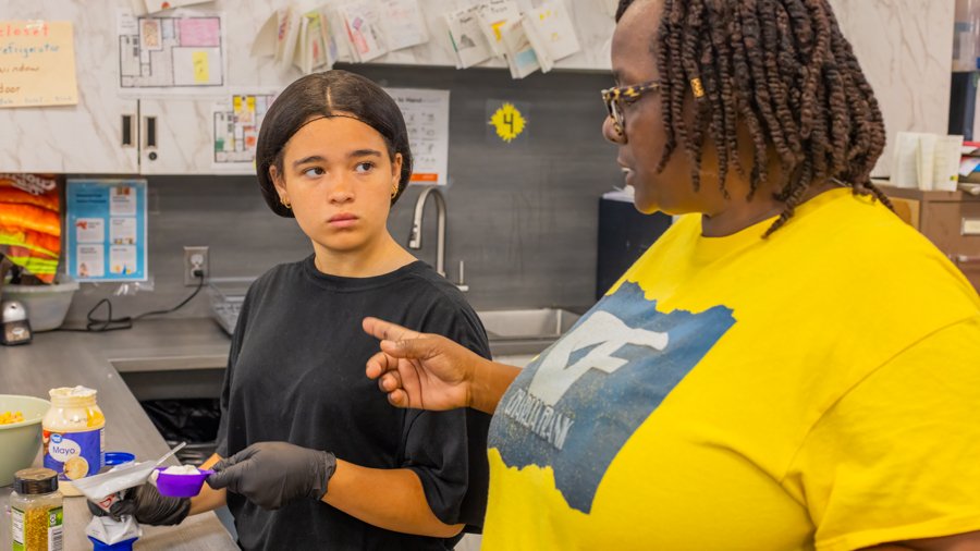 A mentor in a yellow Charlia Frank t-shirt guiding a student who is holding a purple measuring spoon