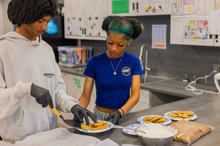 Two teen students carefully plating chocolate chip cookies onto decorative blue-and-white plates with a bowl of whipped cream nearby