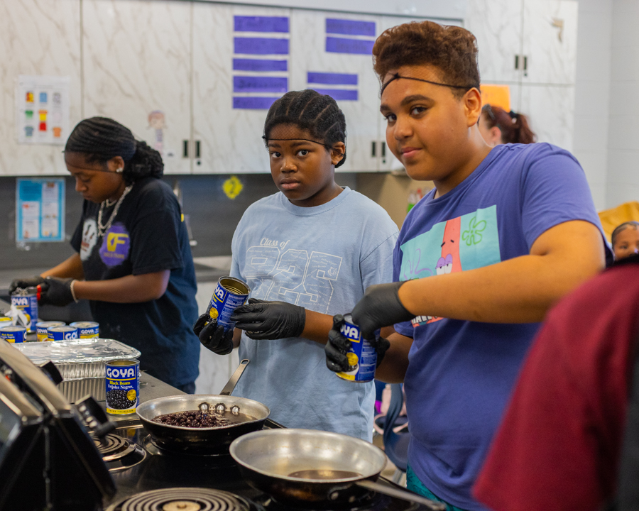 Students working the stovetop together, cooking black beans in pans with cans of beans in hand