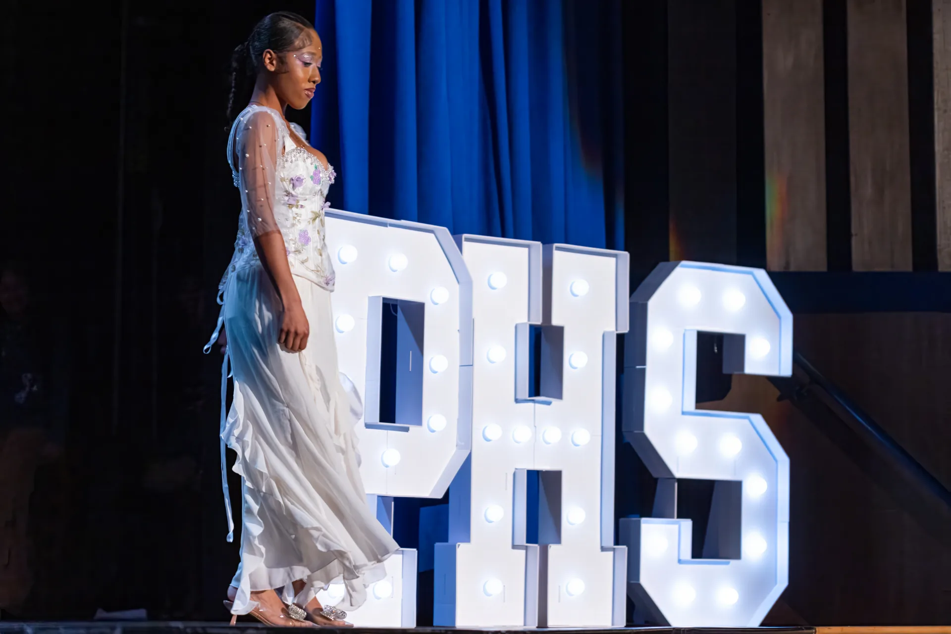 Student in a flowing white formal gown with sheer sleeves standing beside the illuminated PHS letters